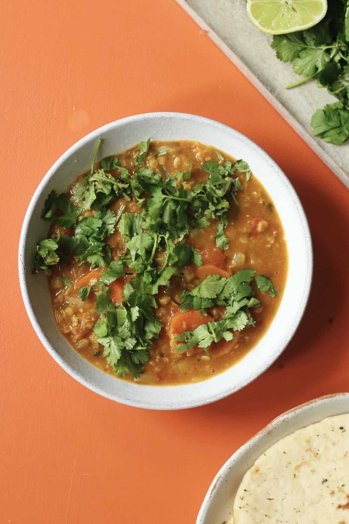 A bowl of red lentil dahl topped with lots of coriander with a separate bowl of naan and coriander and lime.