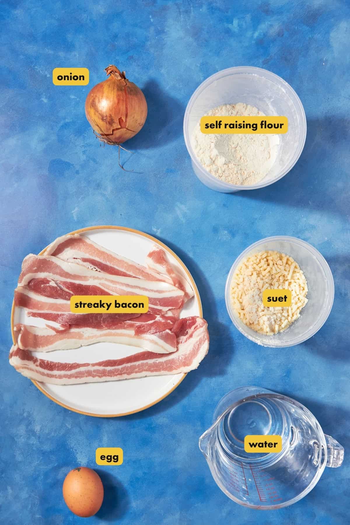 A flat lay of labeled ingredients on a blue surface for bacon pudding: a whole onion, a bowl of self-raising flour, a bowl of suet, a plate of streaky bacon, an egg, and a jug of water.