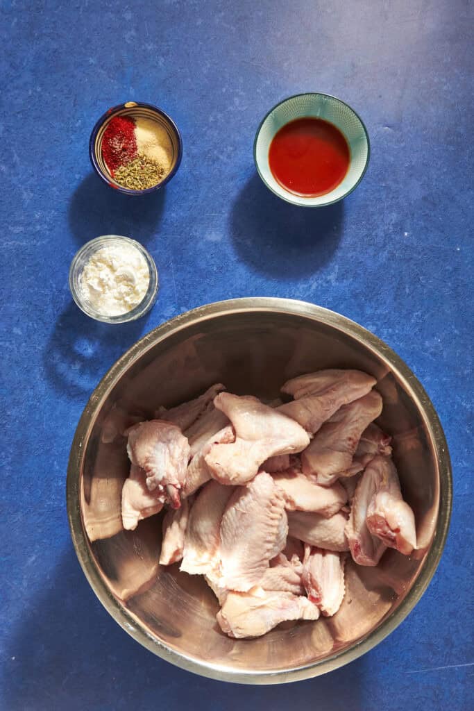 Overhead view of raw chicken wings in a metal bowl, with small bowls of flour, mixed spices, and red hot sauce on a blue countertop—perfect for learning How Long to Bake Chicken Wings at 400 for delicious results.