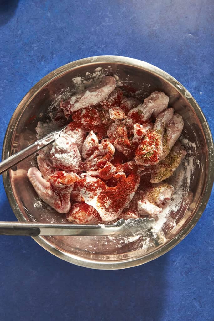 A stainless steel bowl on a blue countertop contains raw chicken wings coated with flour and red spices, ready to be cooked. Wondering how long to bake chicken wings at 400? A pair of metal tongs rests in the bowl, ready for use.