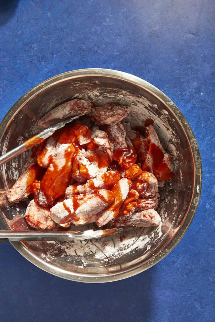 A metal bowl containing floured chicken pieces being coated with red sauce, with a pair of metal tongs resting inside, on a bright blue countertop—perfect prep before discovering how long to bake chicken wings at 400 for crispy results.