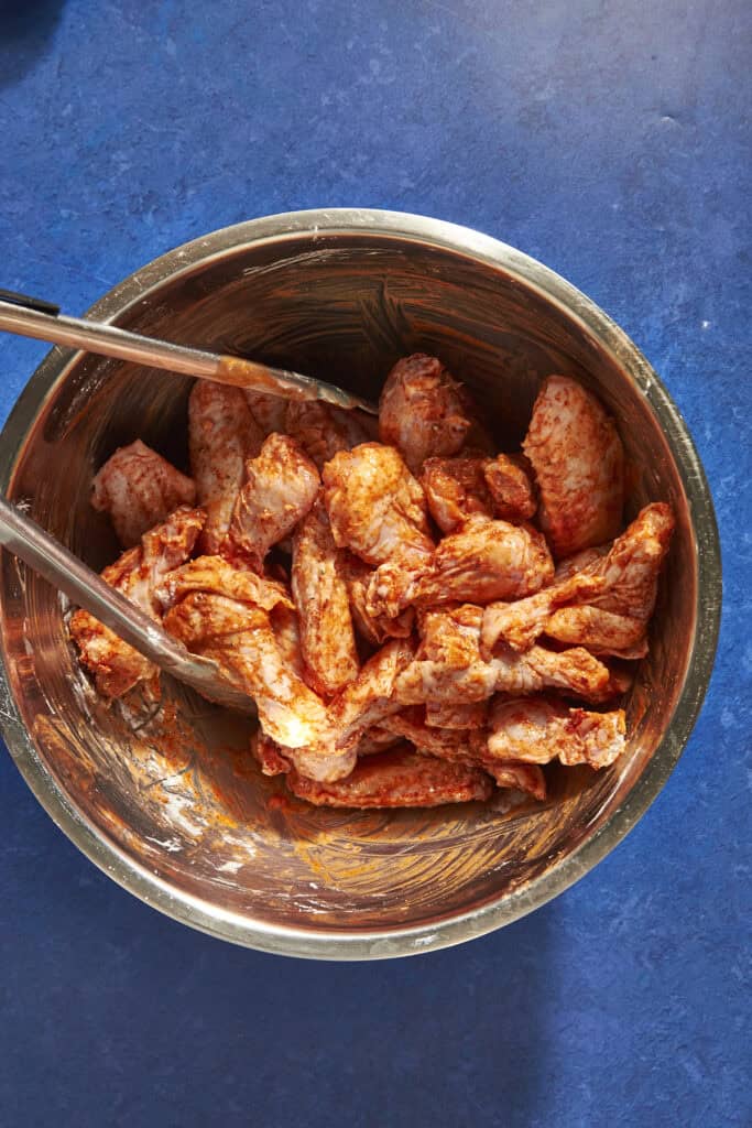 A metal bowl filled with raw chicken wings coated in a red seasoning, with metal tongs resting on the edge, sits on a blue countertop—perfect for those wondering how long to bake chicken wings at 400 for crispy results.