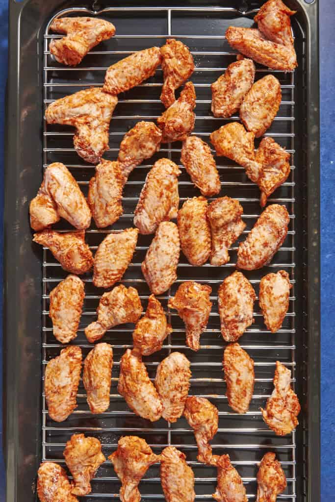 A baking tray lined with a metal rack holds rows of uncooked, seasoned chicken wings spread out evenly, ready for baking. Wondering how long to bake chicken wings at 400? The tray sits on a blue surface.