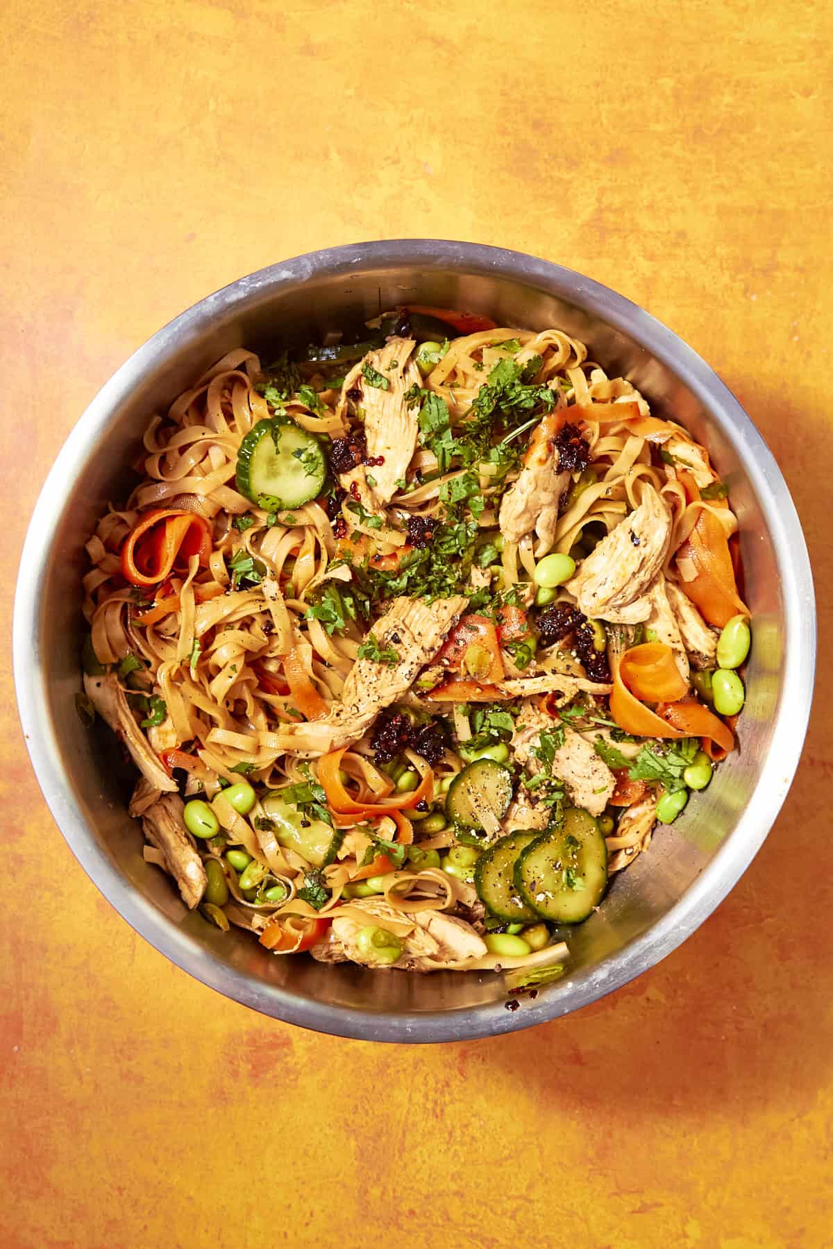 A metal bowl filled with noodle salad featuring shredded chicken, sliced carrots, cucumbers, edamame, roasted corn, and herbs, placed on an orange surface.