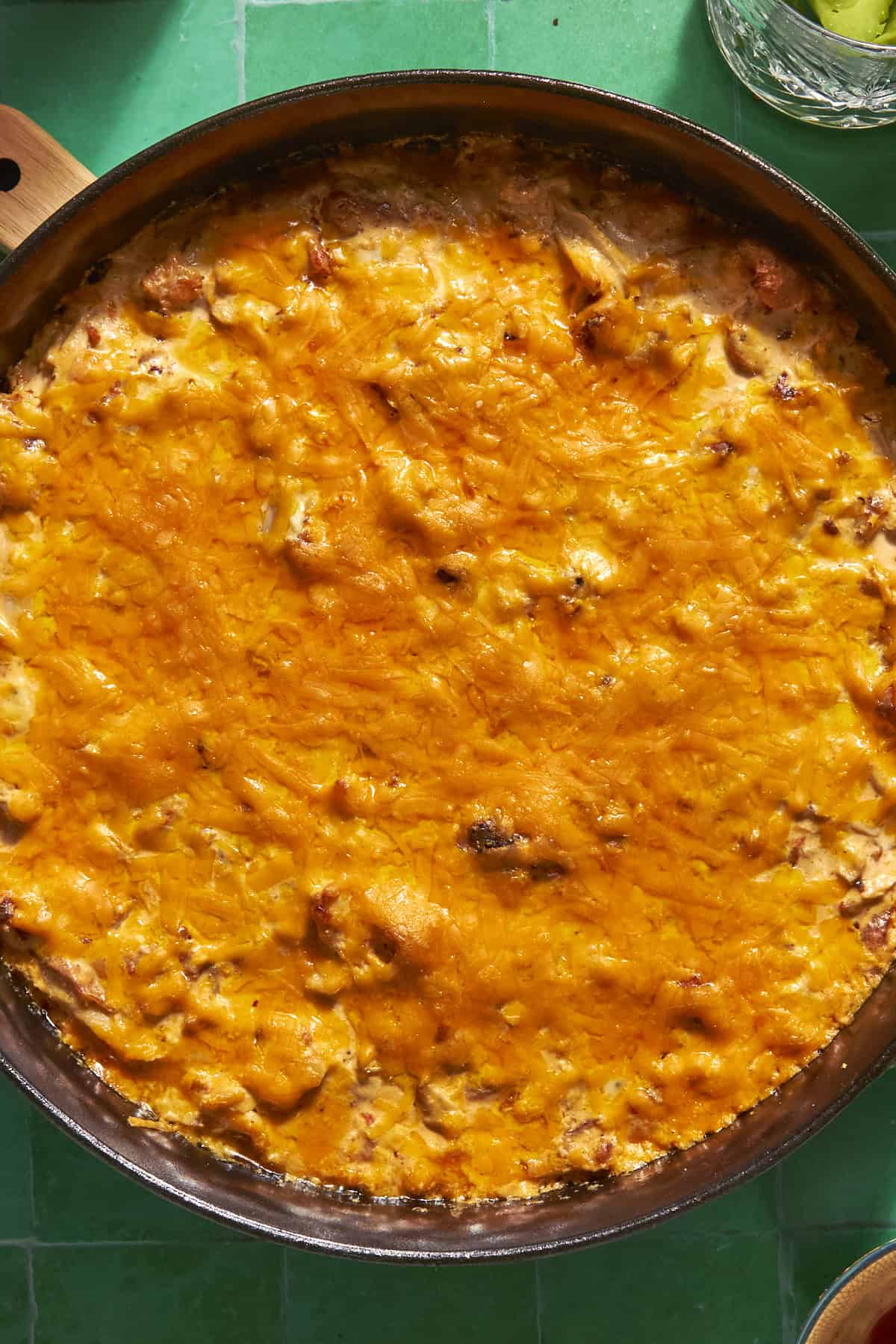 A close-up of a baked casserole dish topped with a golden, melted cheese layer and roasted corn, sitting on a green tiled surface.