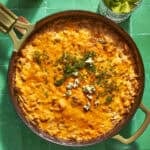 A skillet filled with cheesy, baked dip topped with herbs, crumbled cheese, and roasted corn sits on a green tiled surface, next to a glass with celery sticks and a bottle.
