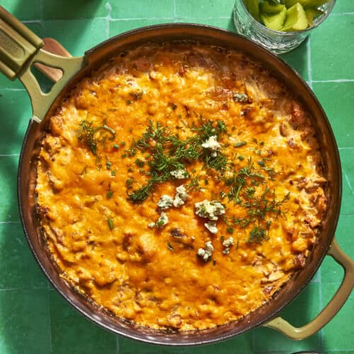 A skillet filled with cheesy, baked dip topped with herbs, crumbled cheese, and roasted corn sits on a green tiled surface, next to a glass with celery sticks and a bottle.