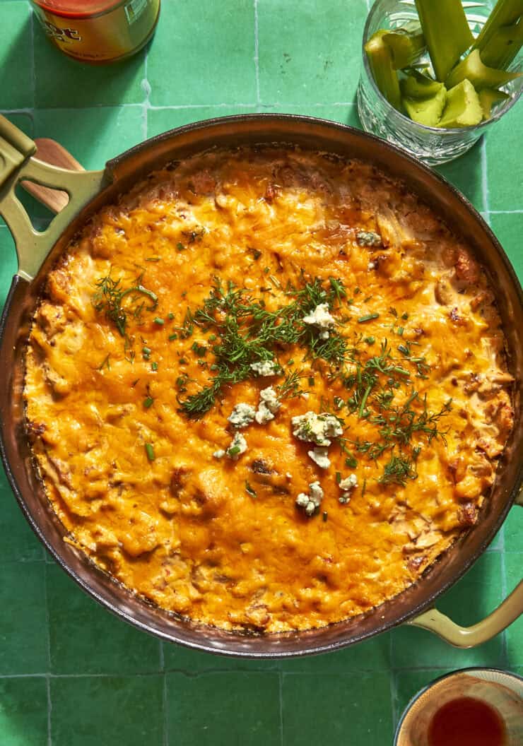 A skillet filled with cheesy, baked dip topped with herbs, crumbled cheese, and roasted corn sits on a green tiled surface, next to a glass with celery sticks and a bottle.