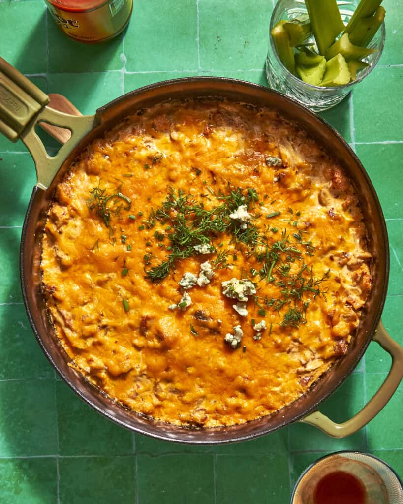 A skillet filled with cheesy, baked dip topped with herbs, crumbled cheese, and roasted corn sits on a green tiled surface, next to a glass with celery sticks and a bottle.