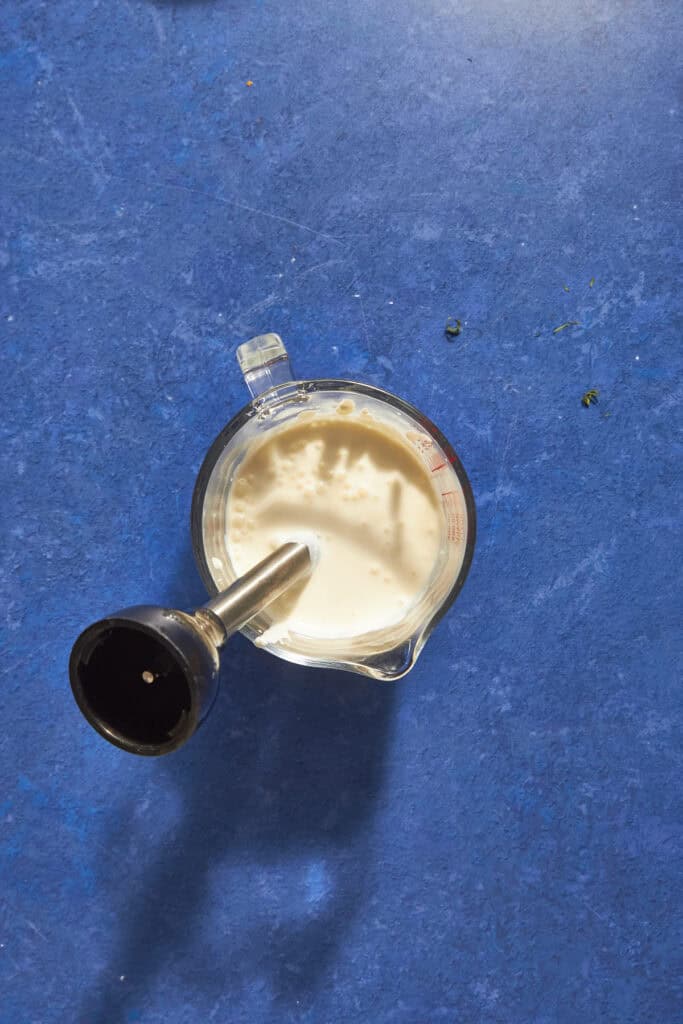 A glass measuring cup filled with creamy roasted corn liquid sits on a blue countertop. An immersion blender is inside the cup, suggesting the mixture is being blended or mixed.