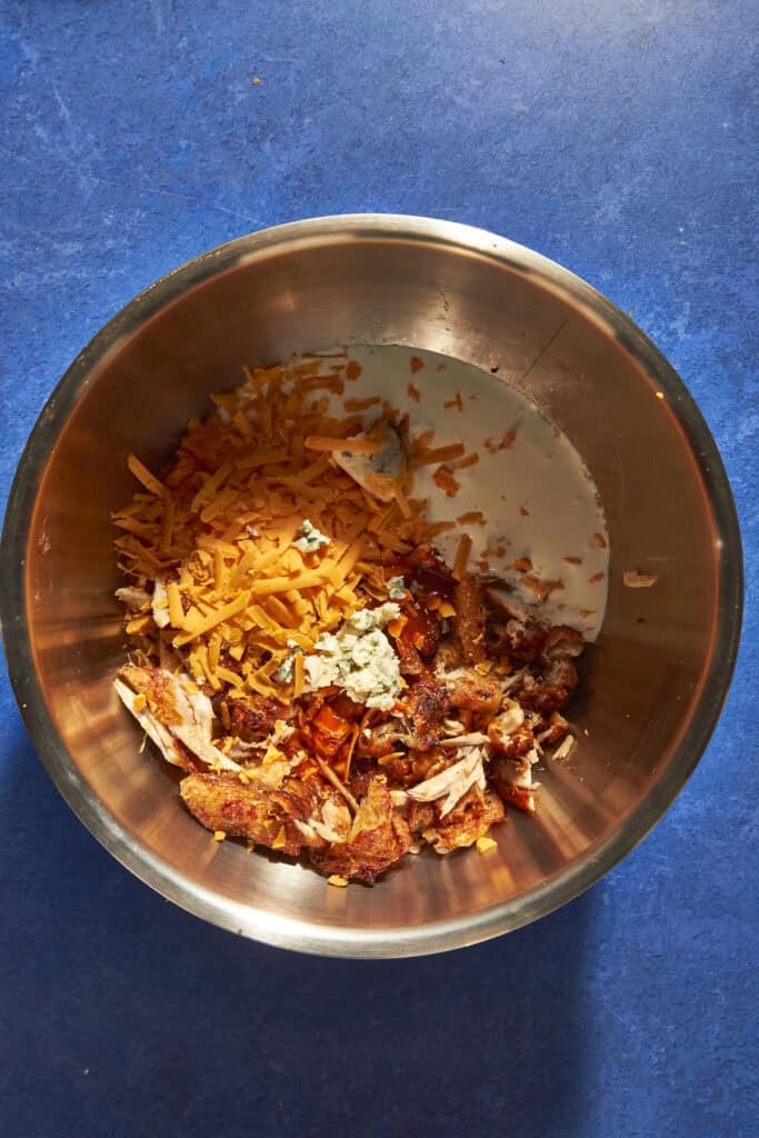 A metal mixing bowl on a blue countertop contains shredded cheddar cheese, chunks of blue cheese, roasted corn, cooked chicken pieces, and a splash of milk.