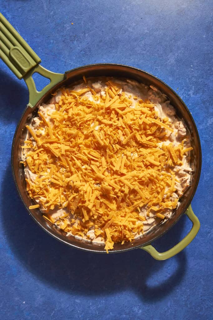 A green-handled skillet on a blue surface filled with a creamy mixture, roasted corn, and topped with a generous layer of shredded cheddar cheese, ready to be cooked or baked.