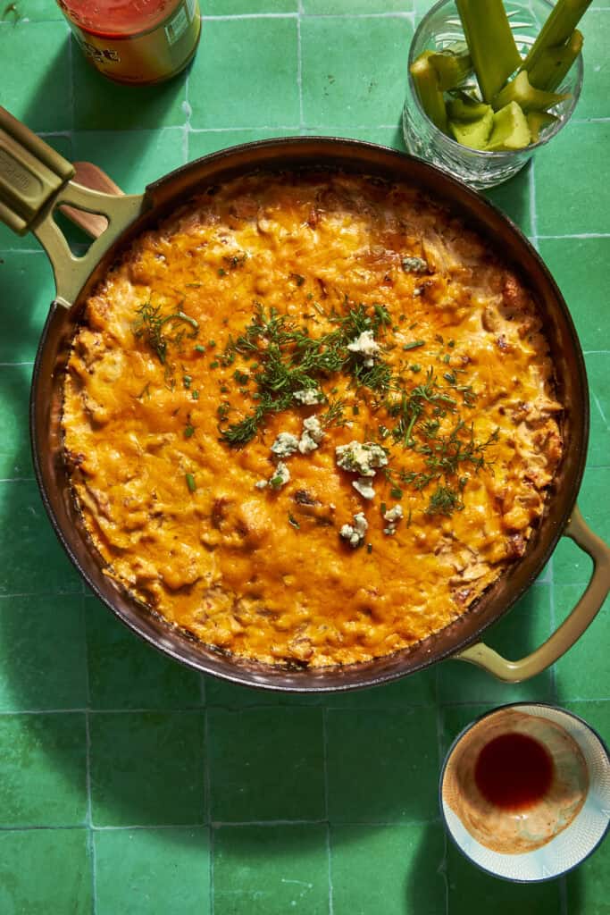 A skillet filled with golden baked cheesy dip and roasted corn, garnished with herbs and blue cheese crumbles, sits on a green tiled surface next to a glass of celery sticks and a small bowl with hot sauce.