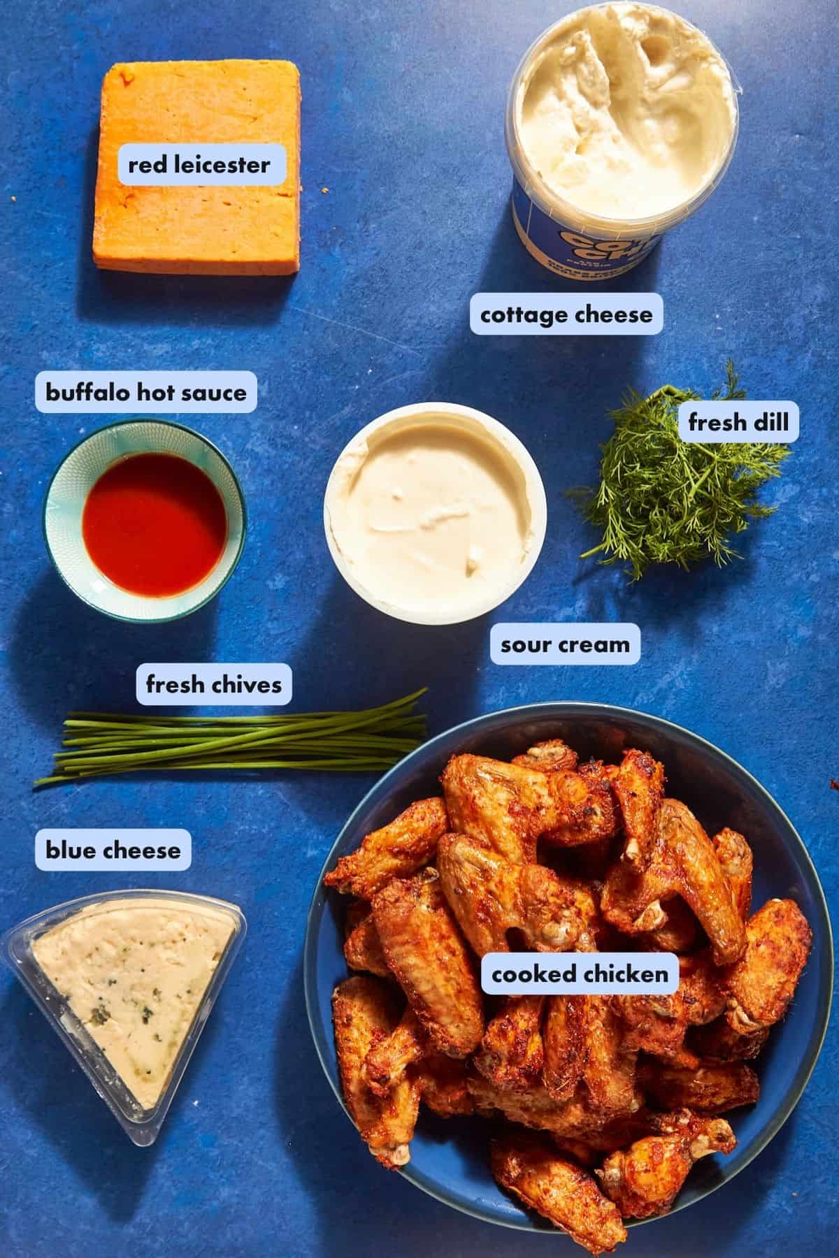 A blue tabletop with labeled ingredients: red leicester cheese, cottage cheese, buffalo hot sauce, fresh chives, blue cheese, sour cream, roasted corn, fresh dill, and a bowl of cooked chicken wings.