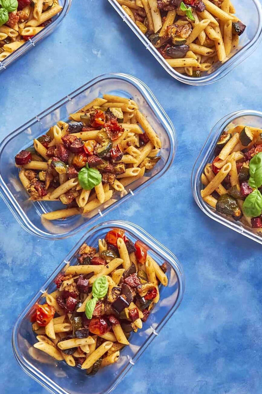 Five clear containers filled with penne pasta, roasted vegetables, and sweet roasted corn, garnished with fresh basil leaves, arranged on a blue background.