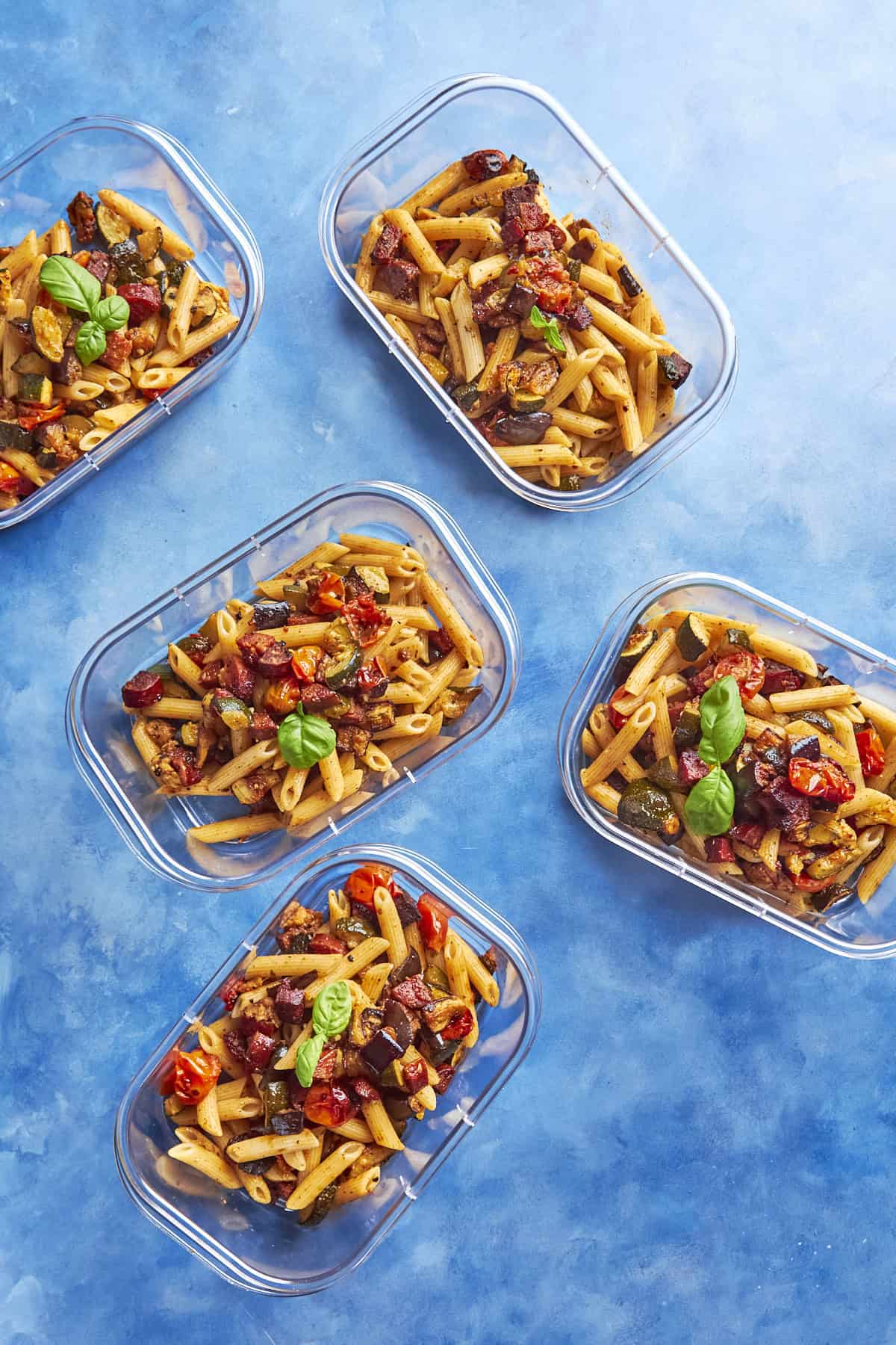 Five clear food containers filled with penne pasta, roasted vegetables, and sweet roasted corn, garnished with fresh basil leaves are arranged on a blue background.