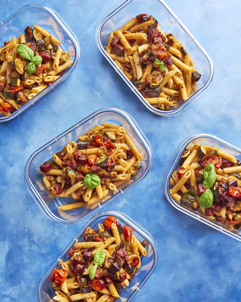 Five clear meal prep containers filled with penne pasta, roasted vegetables, and sweet roasted corn, garnished with basil leaves, arranged on a bright blue background.