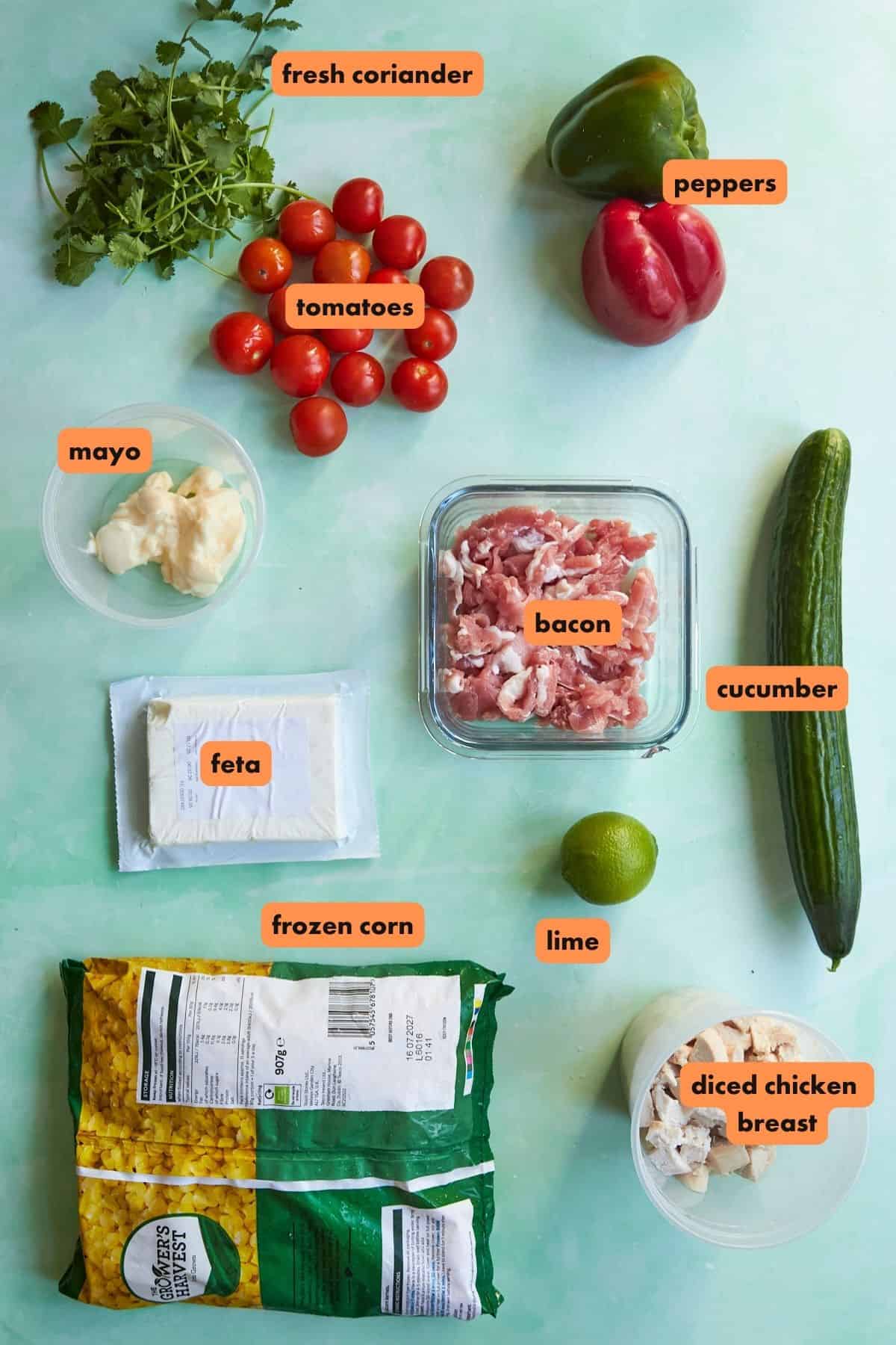 A flat lay of various labeled ingredients on a light green surface: fresh coriander, tomatoes, peppers, mayo, feta, bacon, cucumber, lime, roasted corn, and diced chicken breast.