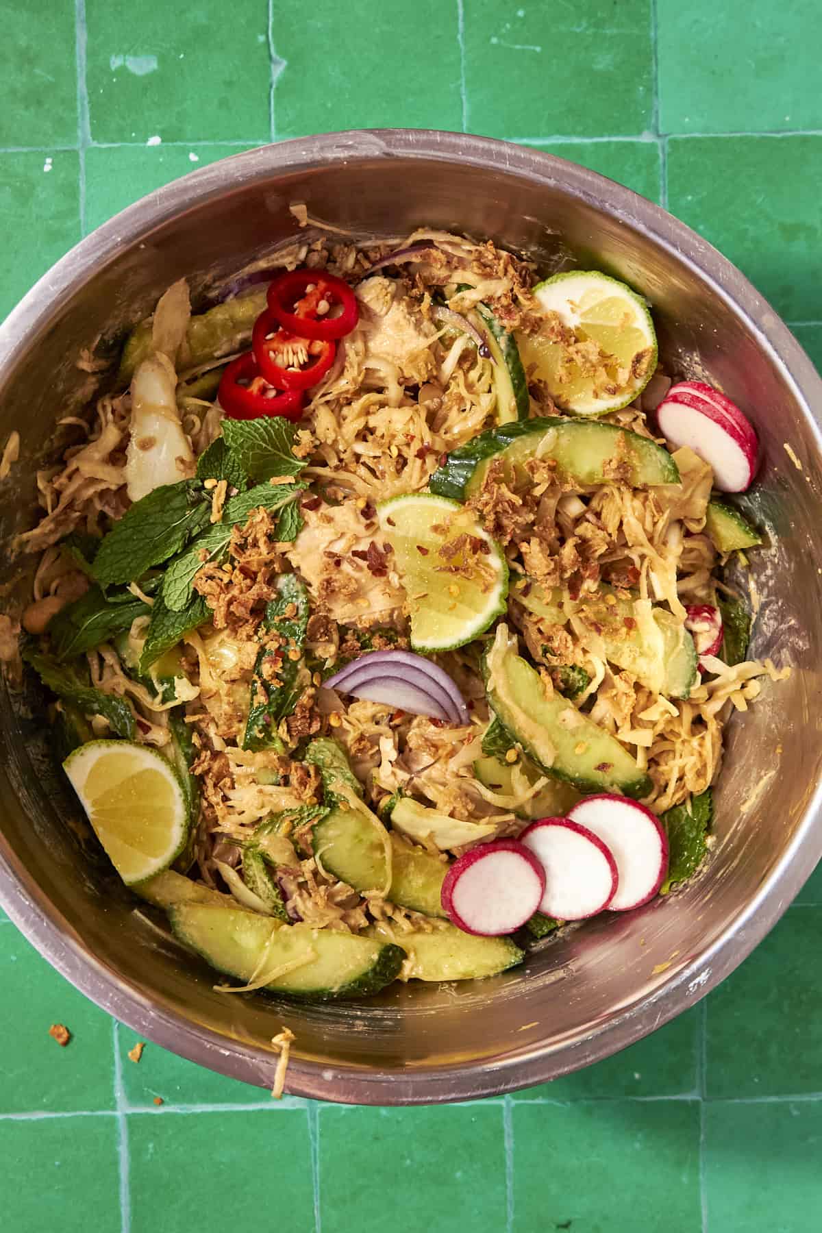 A metal bowl filled with Peanut Chicken Cabbage Salad—shredded cabbage, cucumber, avocado, radish slices, red chili, mint, red onion, crispy toppings, and lime wedges—set on a green tiled surface.
