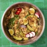 A metal bowl on a green tiled surface filled with Peanut Chicken Cabbage Salad: shredded vegetables, cucumber, radish slices, red chili, lime wedges, mint leaves, and crispy fried onions.