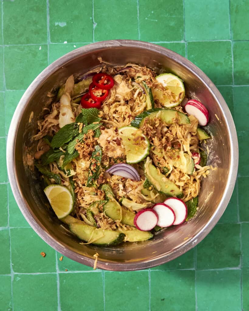 A metal bowl on a green tiled surface filled with Peanut Chicken Cabbage Salad: shredded vegetables, cucumber, radish slices, red chili, lime wedges, mint leaves, and crispy fried onions.