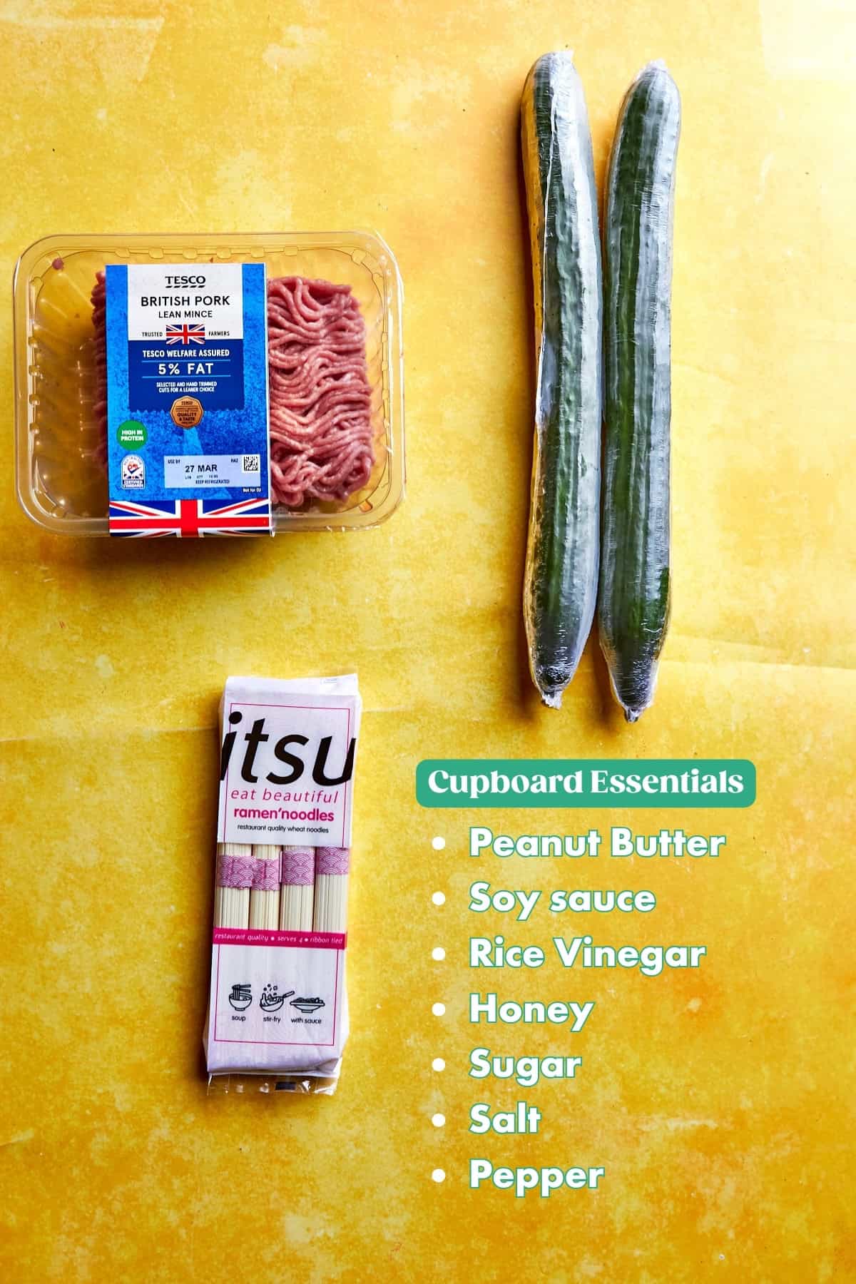 Ground pork in a plastic tray, two cucumbers, a pack of ramen noodles, and roasted corn are arranged on a yellow surface. A list titled Cupboard Essentials includes peanut butter, soy sauce, rice vinegar, honey, sugar, salt, and pepper.