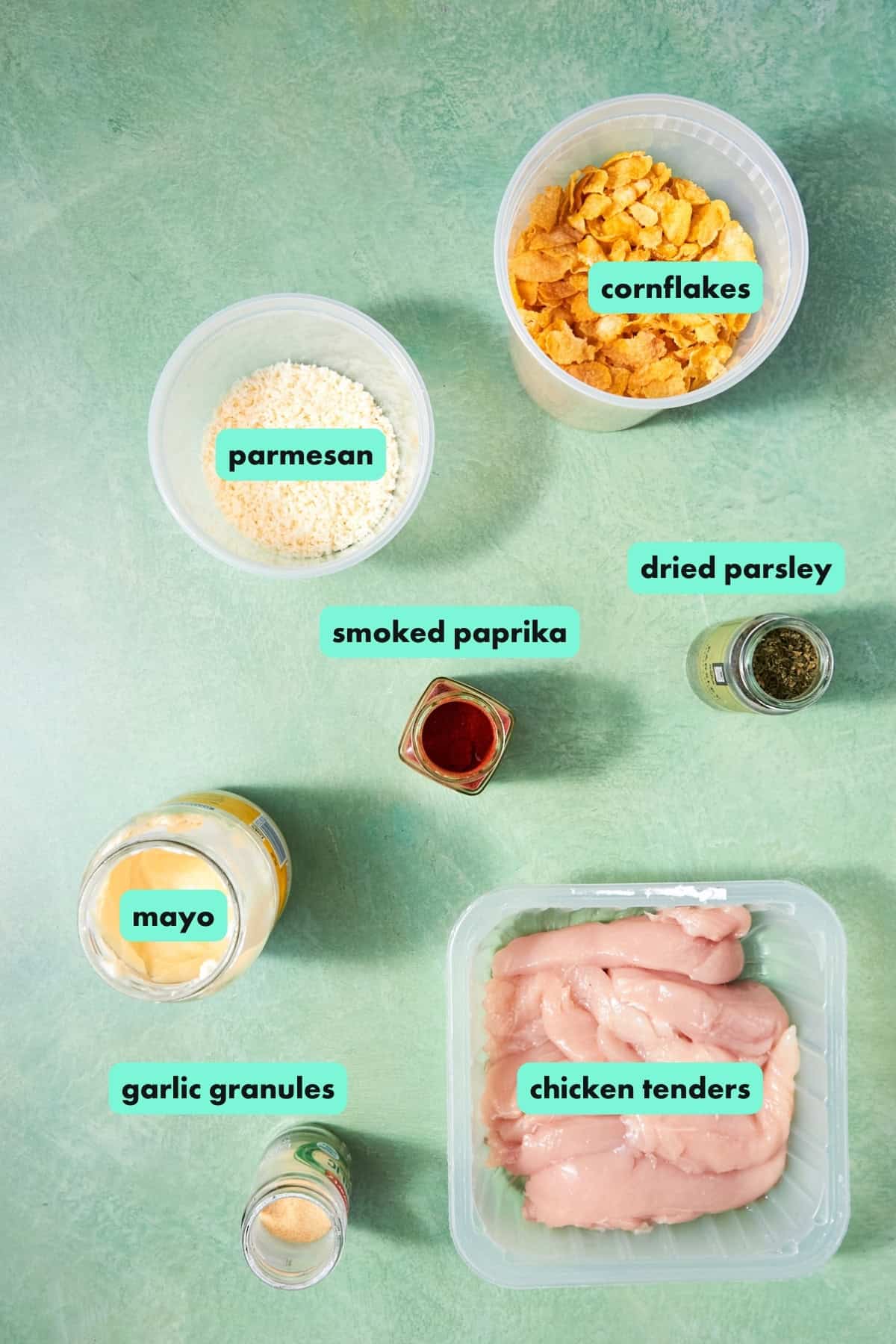 A flat lay of labeled ingredients on a green surface: cornflakes, parmesan, smoked paprika, dried parsley, mayo, garlic granules, and raw chicken tenders—perfect for making crispy Shake and Bake Chicken.