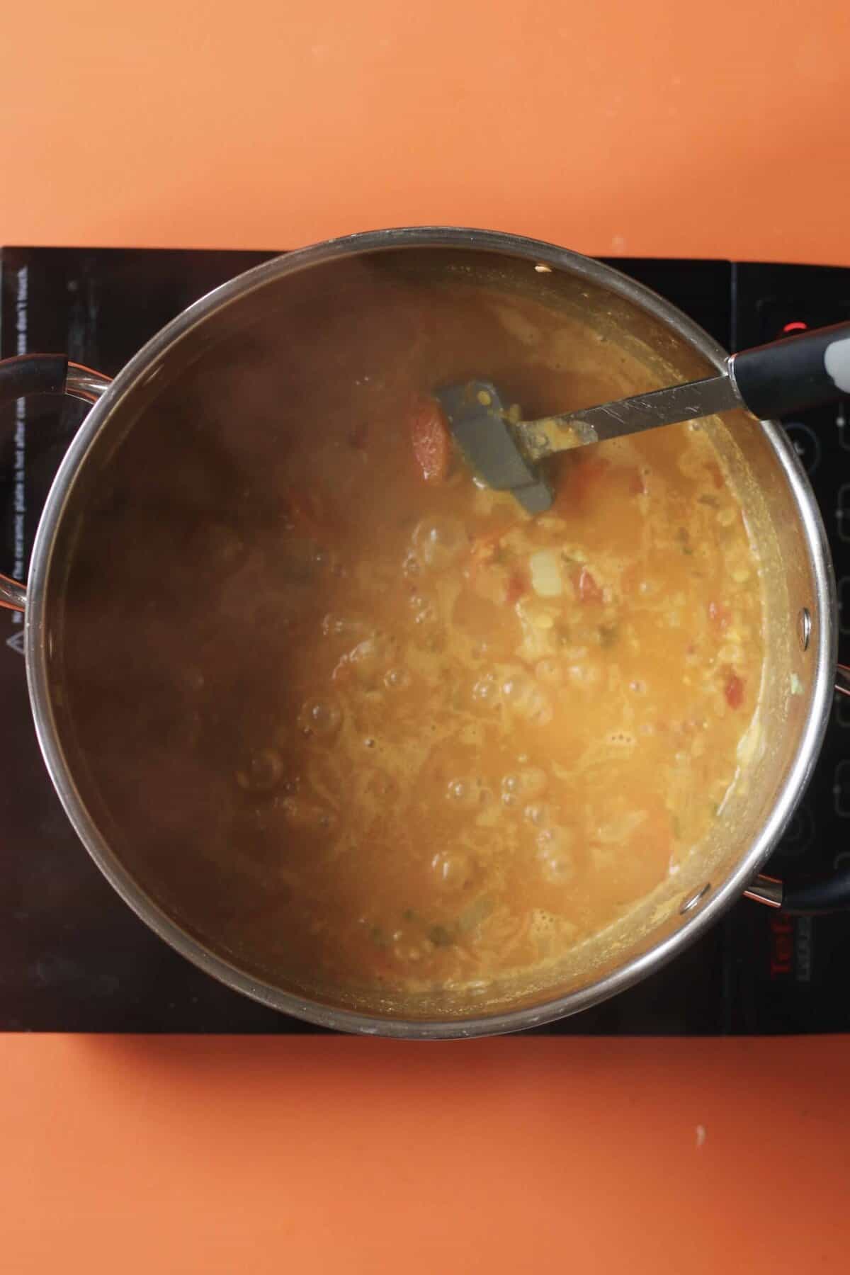 Red lentil dahl cooking a pan and stirred with a spatula on a stove on an orange background.