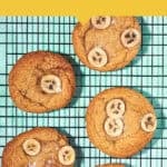 Six banana bread cookies topped with fresh banana slices are cooling on a black wire rack. The teal background and the yellow and white text banners highlight this delicious banana bread cookies recipe and website.