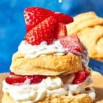 A biscuit shortcake topped with whipped cream and fresh strawberries is stacked on a wooden board, with more shortcake biscuits in the background. Text reads American Shortcake Recipe and Couldn't be easier.