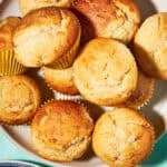A plate of golden-brown Vanilla Muffins, some in yellow paper liners, sits on a turquoise surface with a text overlay: Vanilla Muffins—perfect batch for a crowd, plus a website link at the bottom.