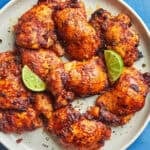 A plate of savory boneless chicken thighs, baked to perfection and garnished with lime wedges, sits on a blue table. Extra lime slices and a baking dish add flair, while text highlights flavor, texture, and a budget-friendly website.