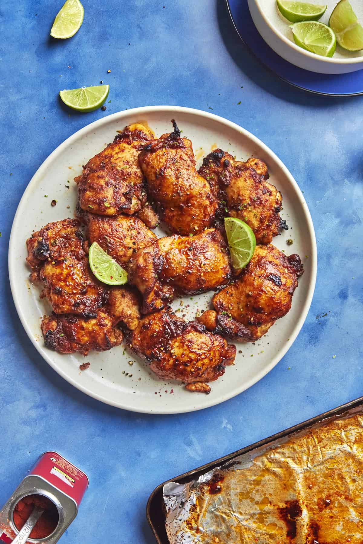 A plate of roasted, seasoned chicken thighs garnished with lime wedges sits on a blue surface. Roasted corn adds color to the dish, while extra lime wedges rest on a small plate nearby, next to part of a baking tray and an open can.