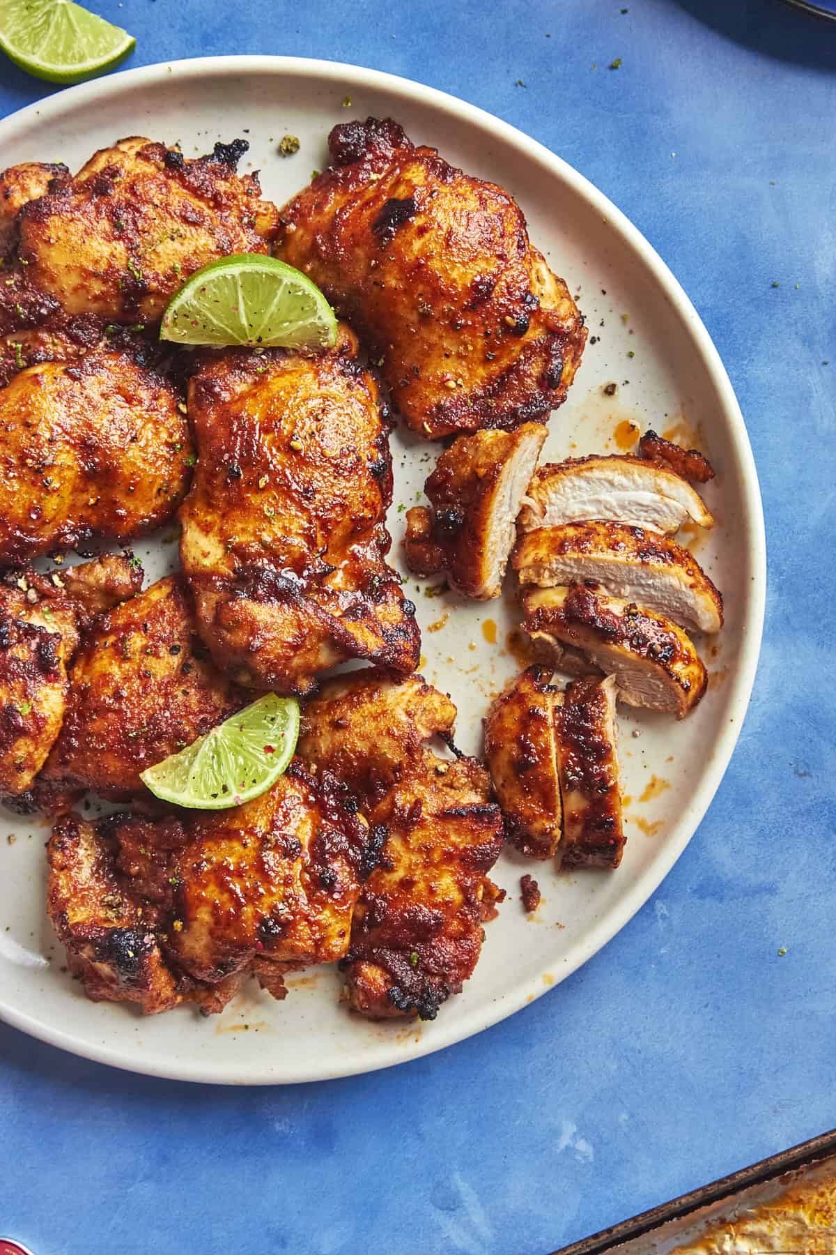 A white plate with several pieces of grilled, seasoned chicken and roasted corn, topped with lime wedges, one piece sliced, on a bright blue background.