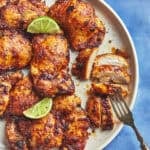 Grilled, seasoned chicken pieces are arranged on a white plate with roasted corn and lime wedges. One piece is sliced, and a fork rests beside it. The plate sits on a blue surface.