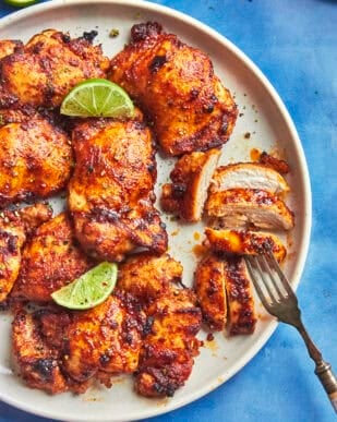 Grilled, seasoned chicken pieces are arranged on a white plate with roasted corn and lime wedges. One piece is sliced, and a fork rests beside it. The plate sits on a blue surface.