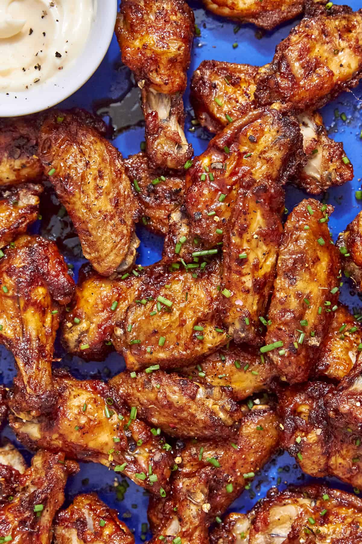 A plate of crispy, seasoned baked chicken wings garnished with chopped chives on a blue surface, with a small bowl of creamy dipping sauce in the corner.