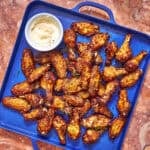 A blue tray filled with baked chicken wings garnished with herbs, accompanied by a small bowl of creamy dipping sauce, sits on a marble surface.