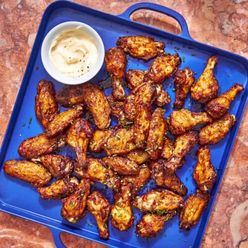 A blue tray filled with baked chicken wings garnished with herbs, accompanied by a small bowl of creamy dipping sauce, sits on a marble surface.