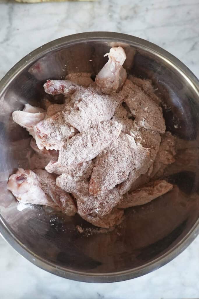 Raw chicken wings, soon to become delicious baked chicken wings, coated with a dry flour and seasoning mixture in a metal mixing bowl on a marble countertop.