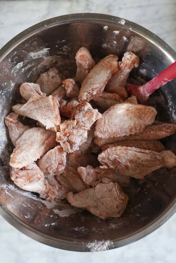 A metal mixing bowl filled with raw baked chicken wings coated in flour and seasoning, with a red spatula partially visible on the right side.