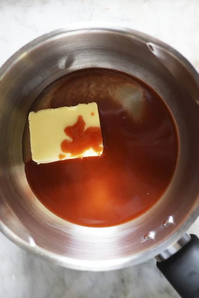A stainless steel saucepan containing a block of butter and a pool of reddish-brown sauce, perfect for tossing baked chicken wings, sits on a light-colored countertop.