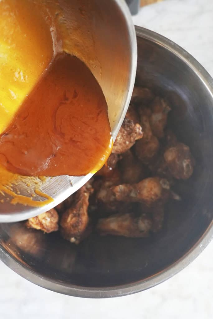 A metal bowl pours orange buffalo sauce over a large bowl filled with baked chicken wings, ready to be coated.