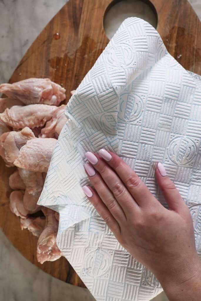 A hand with light pink nail polish presses a paper towel onto raw chicken wings on a wooden cutting board, drying the meat before preparing baked chicken wings.