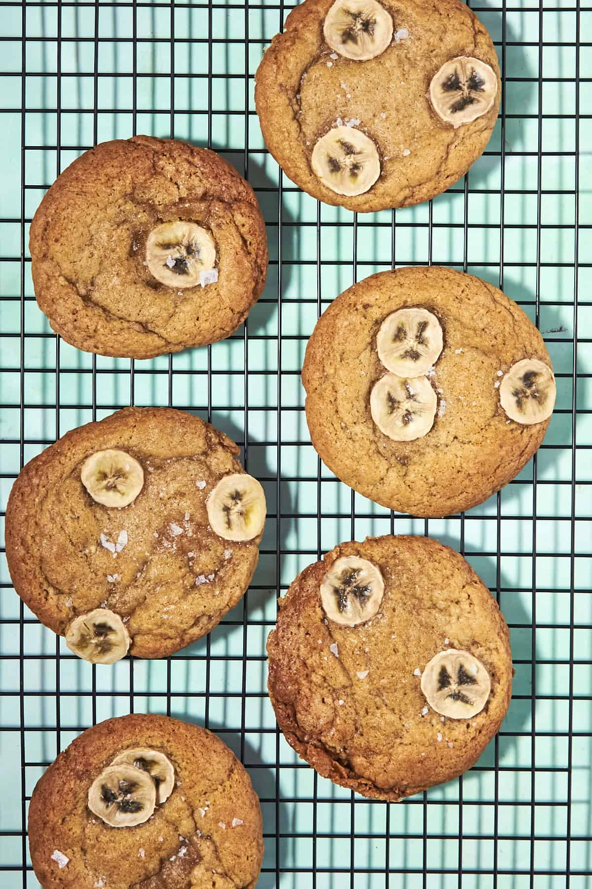 Six golden-brown banana bread cookies topped with fresh banana slices are cooling on a black wire rack over a light blue background.