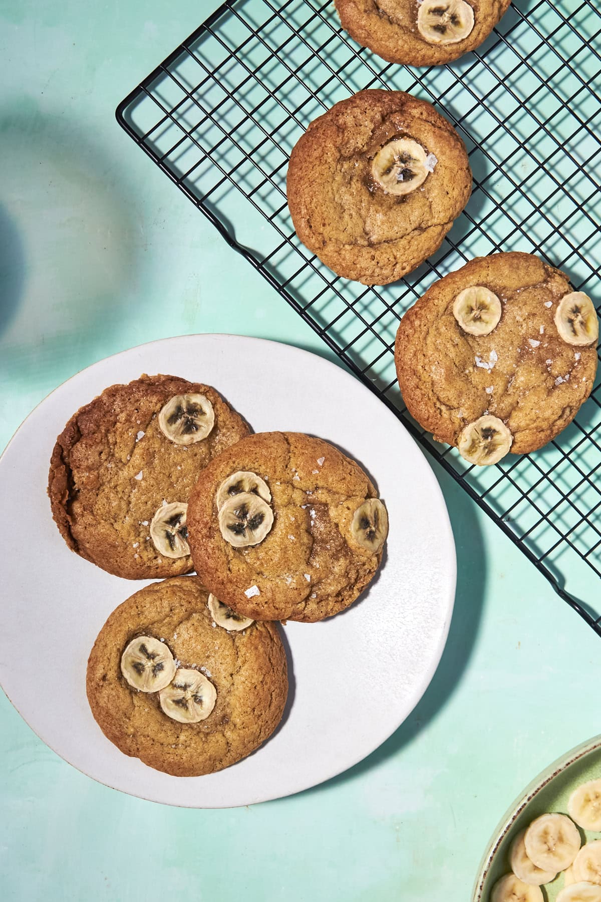 Three banana bread cookies topped with banana slices rest on a white plate beside four more on a black cooling rack, all set on a light turquoise surface, with extra banana slices in a bowl at the corner.