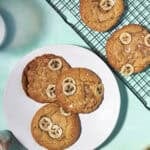 Three banana bread cookies topped with banana slices sit on a white plate, next to two similar cookies cooling on a rack, all arranged on a light blue surface.
