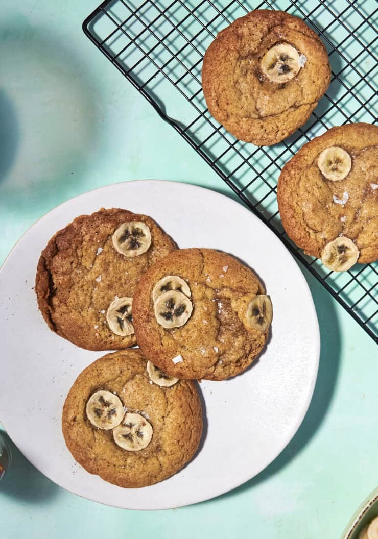 Three banana bread cookies topped with banana slices sit on a white plate, next to two similar cookies cooling on a rack, all arranged on a light blue surface.