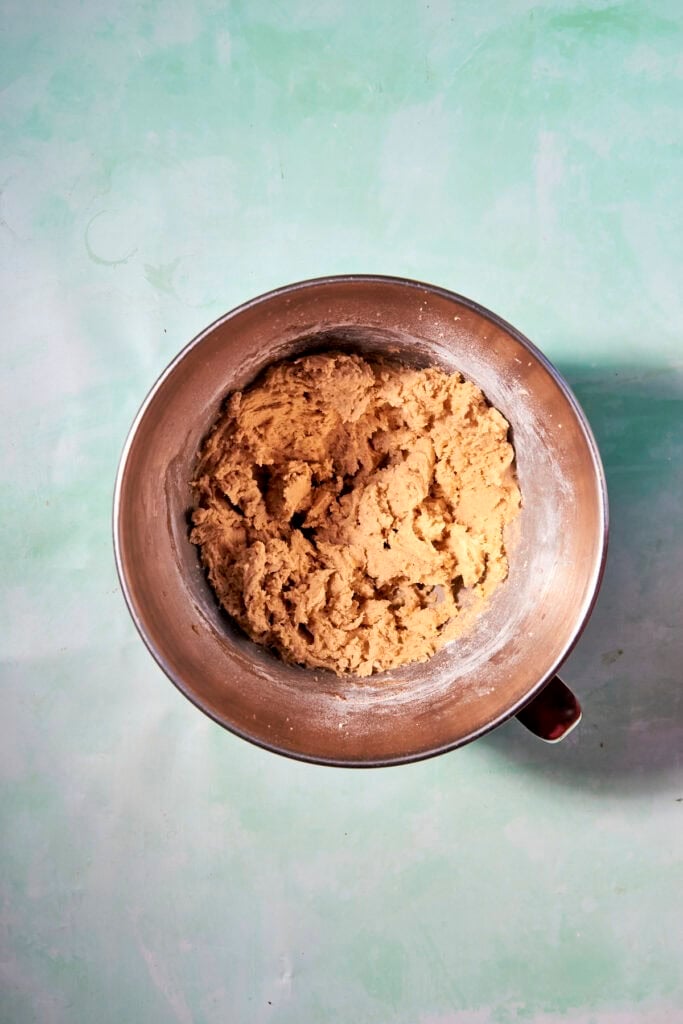 A stainless steel mixing bowl filled with banana bread cookie dough sits on a light green, textured surface viewed from above.