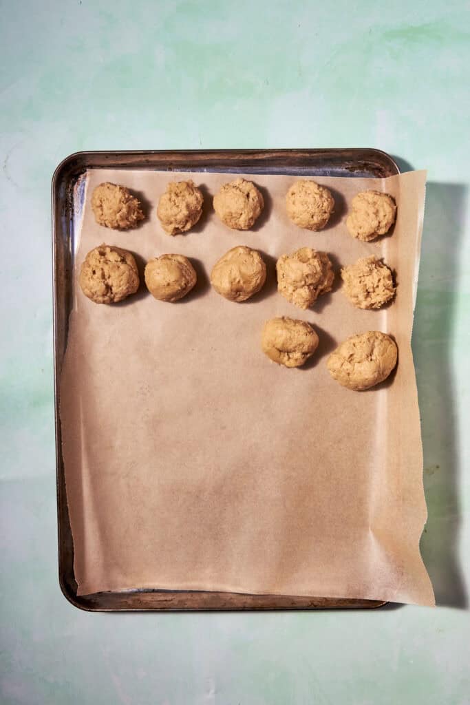 A baking tray lined with parchment paper holds eleven unbaked banana bread cookie dough balls arranged in rows on a greenish background.